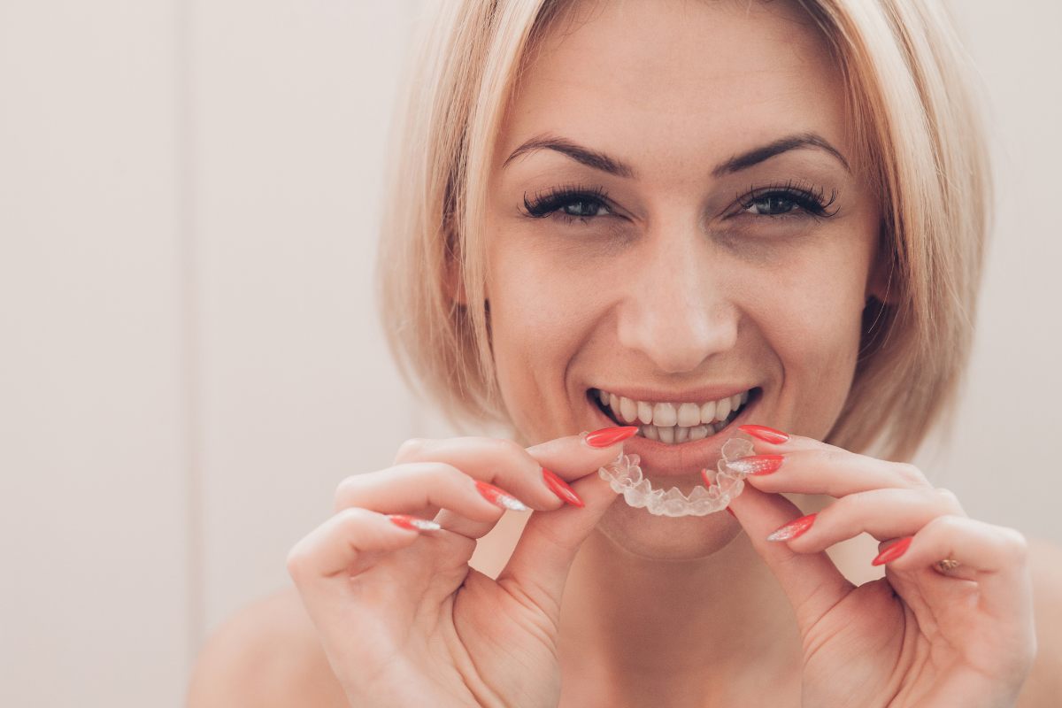 Woman smiling while holding clear dental aligners against her teeth indoors.